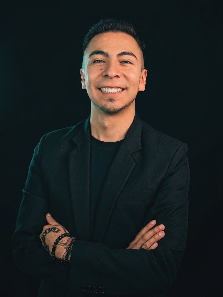 Retrato de Jhonatan Ascárraga, fundador y CEO de Business Development Agency LLC: blazer y camiseta negros sobre fondo negro, brazos cruzados y tatuajes visibles en manos y muñeca.